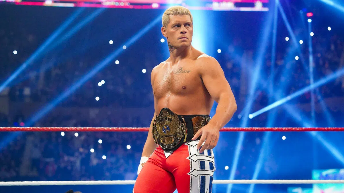 WWE Superstar Cody Rhodes stands confidently in a wrestling ring under bright blue lights, wearing red wrestling tights and holding the historic "Winged Eagle" WWE Championship belt around his waist.