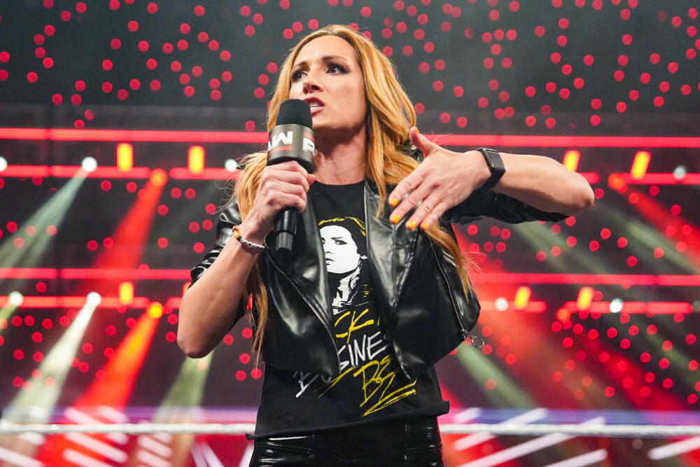 A professional wrestler with long blonde hair stands on a stage holding a black microphone with white “NXT” branding, wearing a black leather vest over a graphic T‑shirt and black pants, with red and white spotlights behind her.