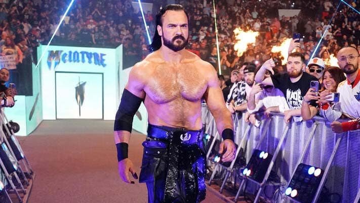 Professional wrestler Drew McIntyre makes his entrance into a crowded arena, walking confidently toward the ring with pyrotechnics firing in the background.