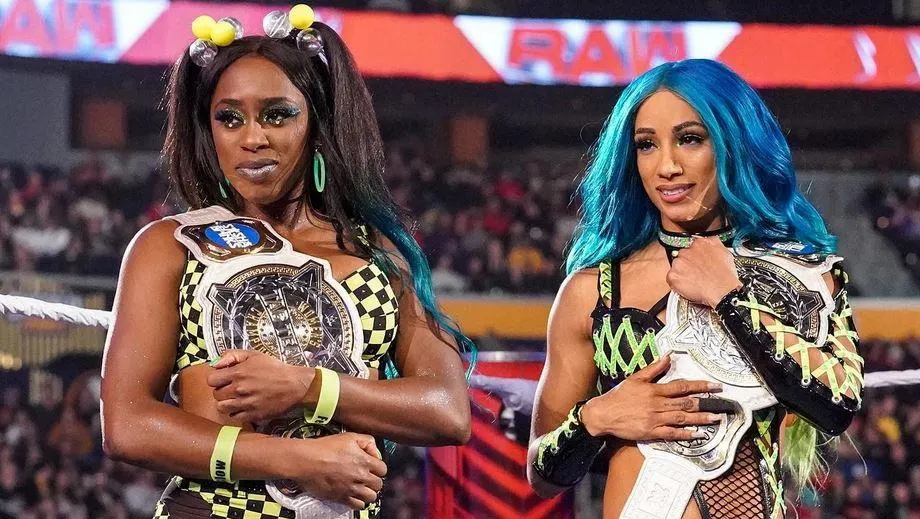 Two female professional wrestlers stand inside a WWE ring holding championship belts over their shoulders during a live televised event.