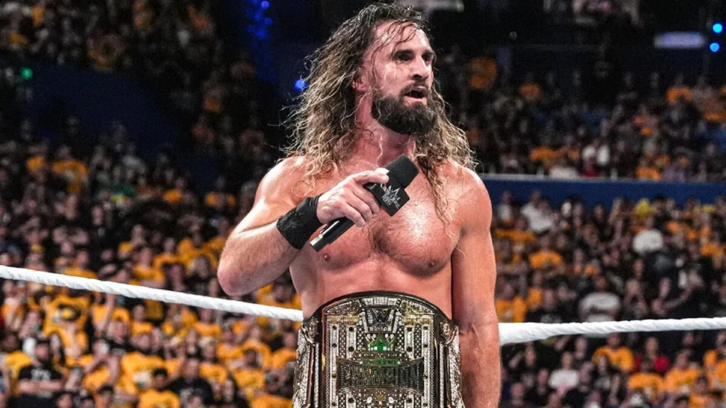 A shirtless professional wrestler with long, wet hair and a beard stands in a wrestling ring holding a microphone in his right hand and a large silver championship belt in front of him, with a crowd of spectators wearing yellow in the background.