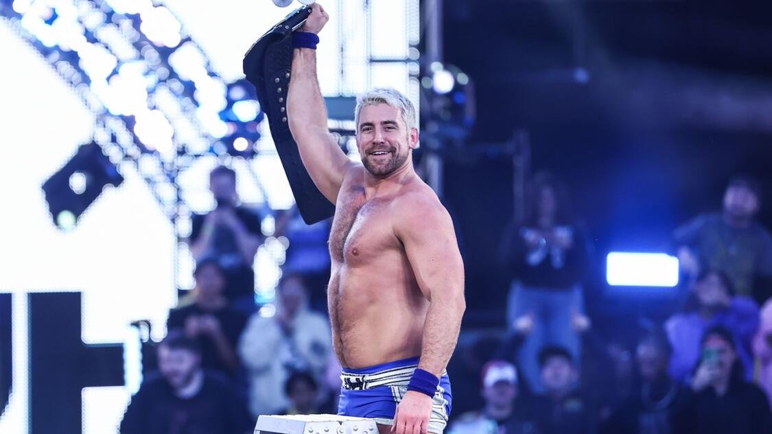 Professional wrestler Joe Hendry stands confidently in the ring with his arms raised, holding the NXT Championship belt over his shoulder. He has short, light brown hair and a beard, and is wearing blue and white wrestling gear.