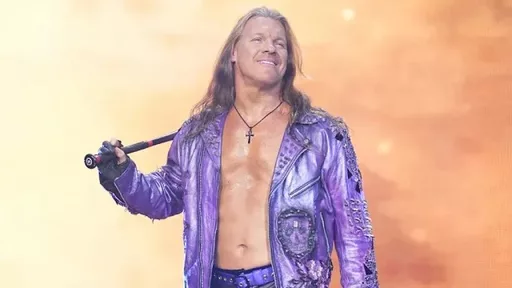 Professional wrestler Chris Jericho, with long hair and a purple sparkly jacket, smiles in a brightly lit arena.