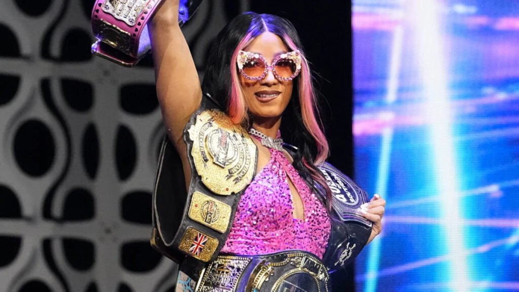 Professional wrestler Mercedes Moné stands triumphantly in an AEW ring, smiling while wearing cat-eye glasses and holding multiple championship belts. She is dressed in a sparkling pink sleeveless outfit with her signature pink-streaked dark hair.