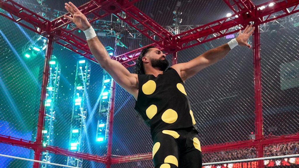 Seth Rollins stands inside a red steel cage with his arms raised, wearing black gear with large yellow polka dots during a WWE match.