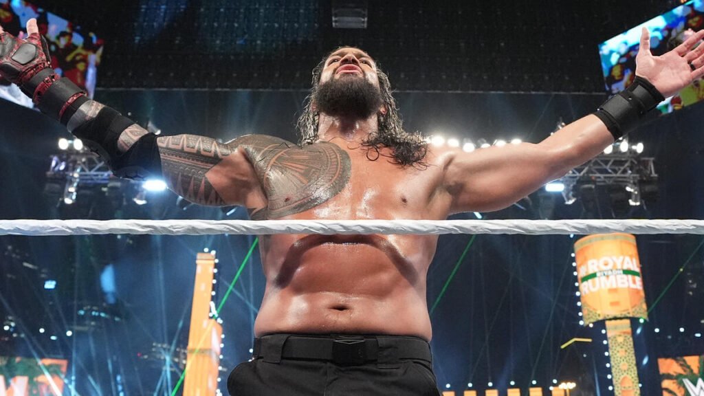 Professional wrestler Roman Reigns, with long dark hair and a beard, stands in the ring with his arms spread wide in a triumphant pose.