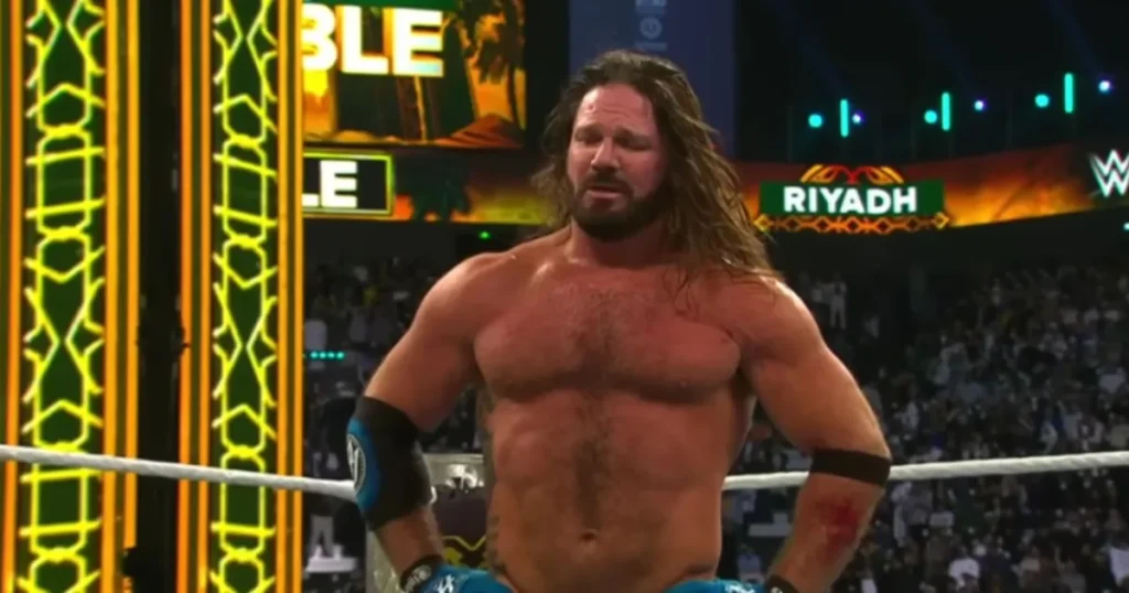 A close-up of professional wrestler AJ Styles standing in a wrestling ring, looking exhausted and emotional. He has long, sweat-drenched brown hair and a beard. He is shirtless, showing his athletic build and a large tattoo on his right side. He is wearing blue and black wrestling trunks and padded elbow sleeves. The background shows a brightly lit arena with a "RIYADH" sign and the blurred faces of a large crowd.