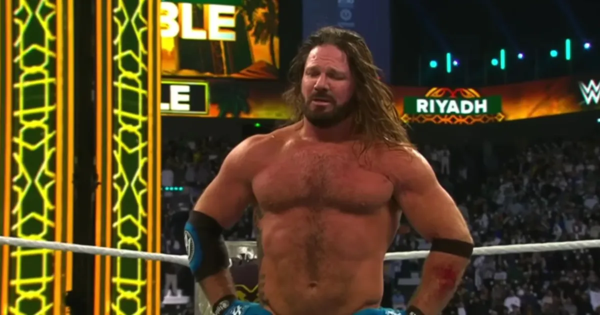A close-up of professional wrestler AJ Styles standing in a wrestling ring, looking exhausted and emotional. He has long, sweat-drenched brown hair and a beard. He is shirtless, showing his athletic build and a large tattoo on his right side. He is wearing blue and black wrestling trunks and padded elbow sleeves. The background shows a brightly lit arena with a "RIYADH" sign and the blurred faces of a large crowd.