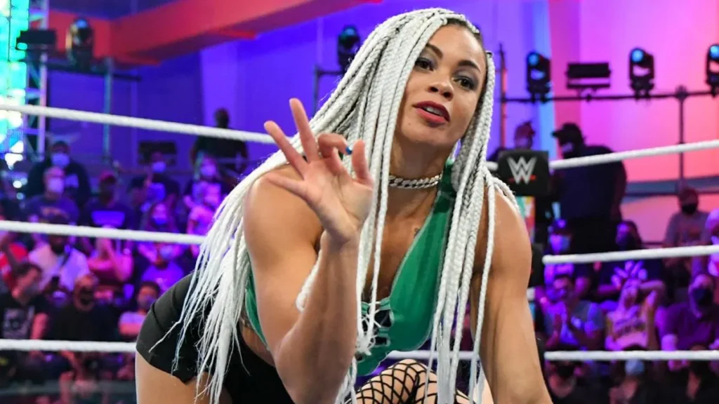 B-Fab, a professional wrestler with long white braids, poses in a green top and black shorts in a brightly lit wrestling ring.