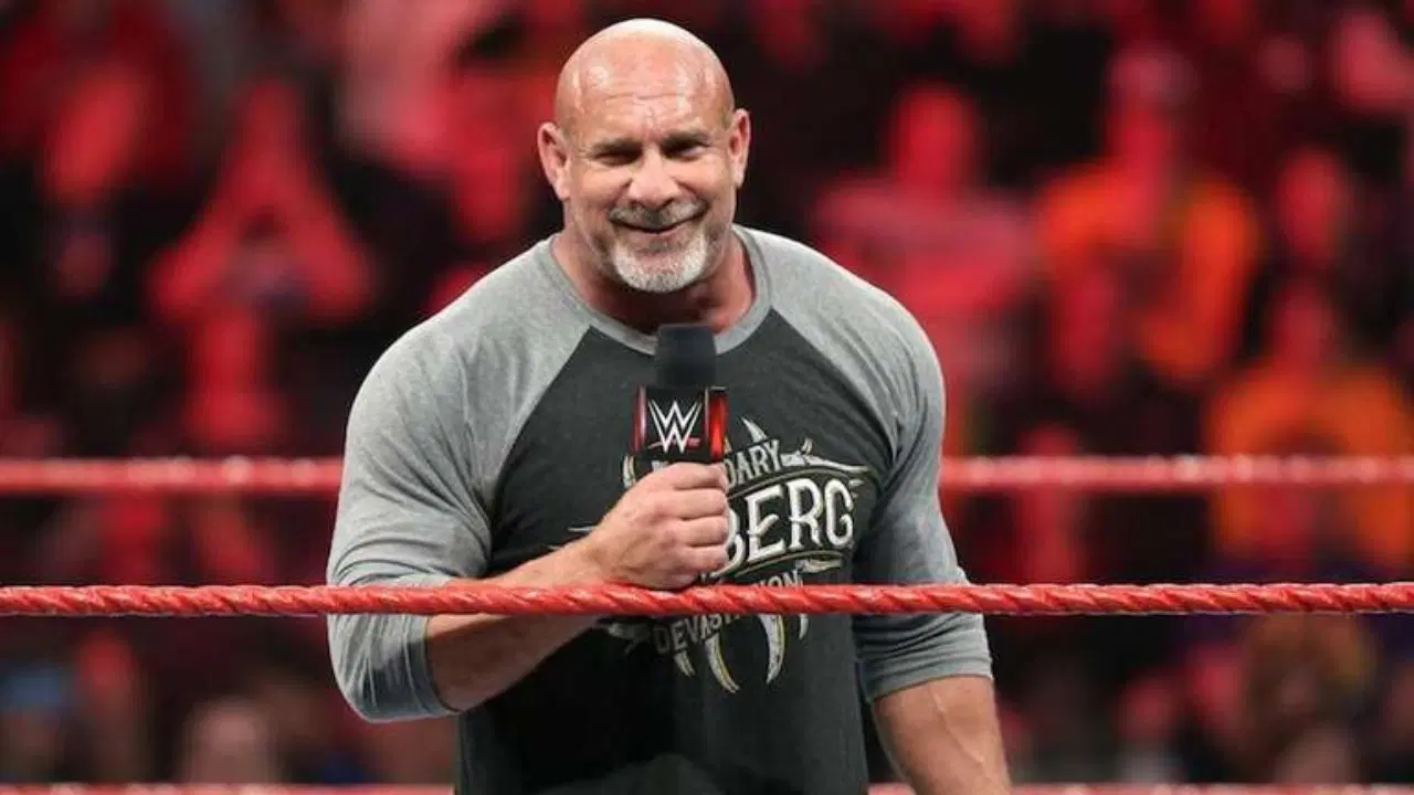A close-up of professional wrestler Goldberg standing in a wrestling ring, smiling and holding a WWE-branded microphone. He is bald with a grey-flecked beard and is wearing a black and grey baseball-style T-shirt with "GOLDBERG" and the word "LEGENDARY" printed on it. The background shows the blurred red and black colors of a packed arena crowd.