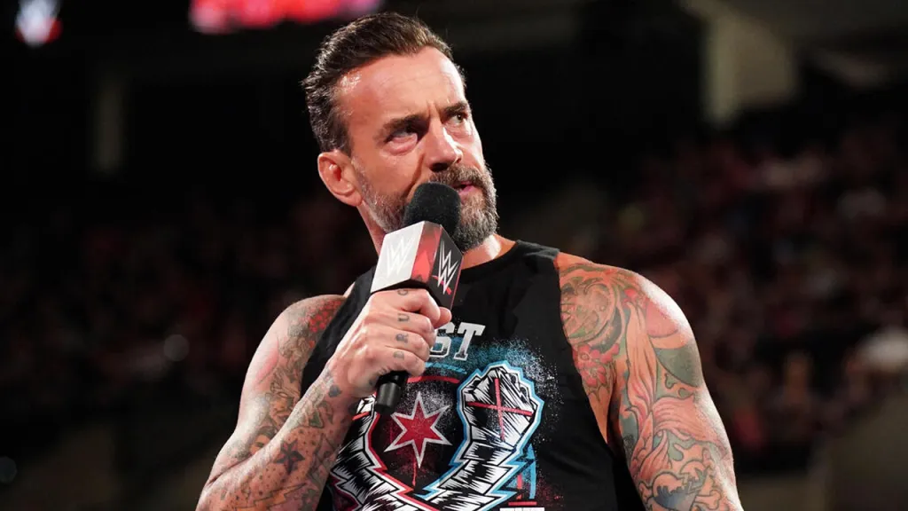 A close-up of professional wrestler CM Punk standing in a wrestling ring, holding a WWE-branded microphone and looking off-camera with a serious expression. He has short dark hair, a salt-and-pepper beard, and extensive colorful tattoos covering both arms and his shoulders. He is wearing a black sleeveless shirt featuring his "Best in the World" logo.