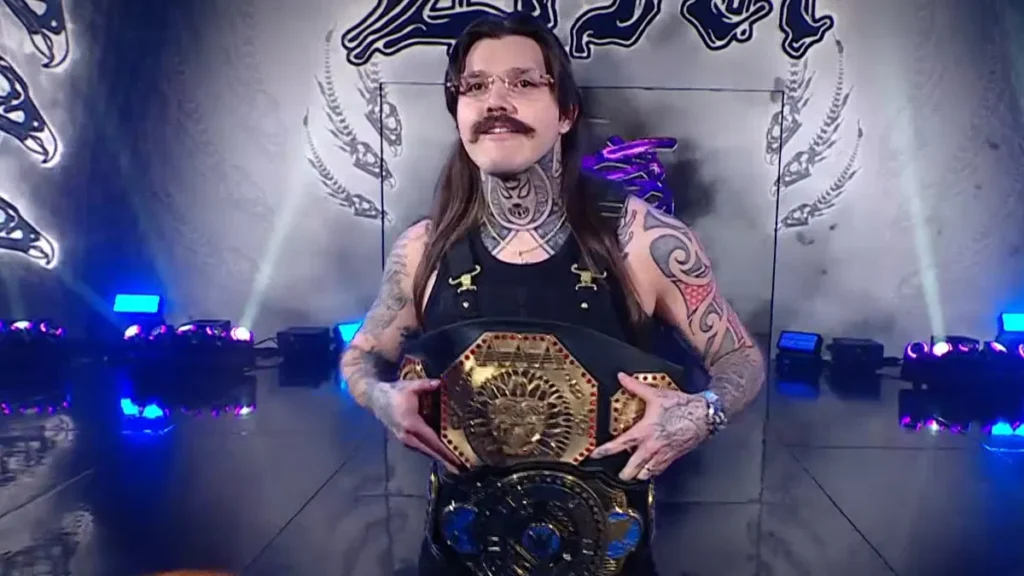 Professional wrestler Dominik Mysterio, with a mustache, glasses, and tattoos, poses while holding two championship belts in a ring.