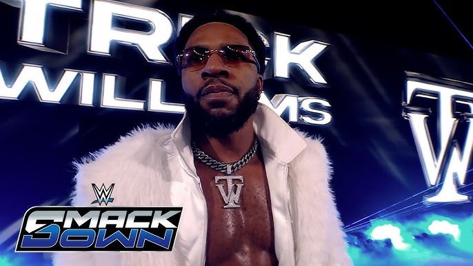 Professional wrestler Trick Williams stands in the center of a WWE SmackDown entrance, wearing a white fur-collared jacket and a silver "TW" pendant, with his name and logo displayed on large screens behind him.