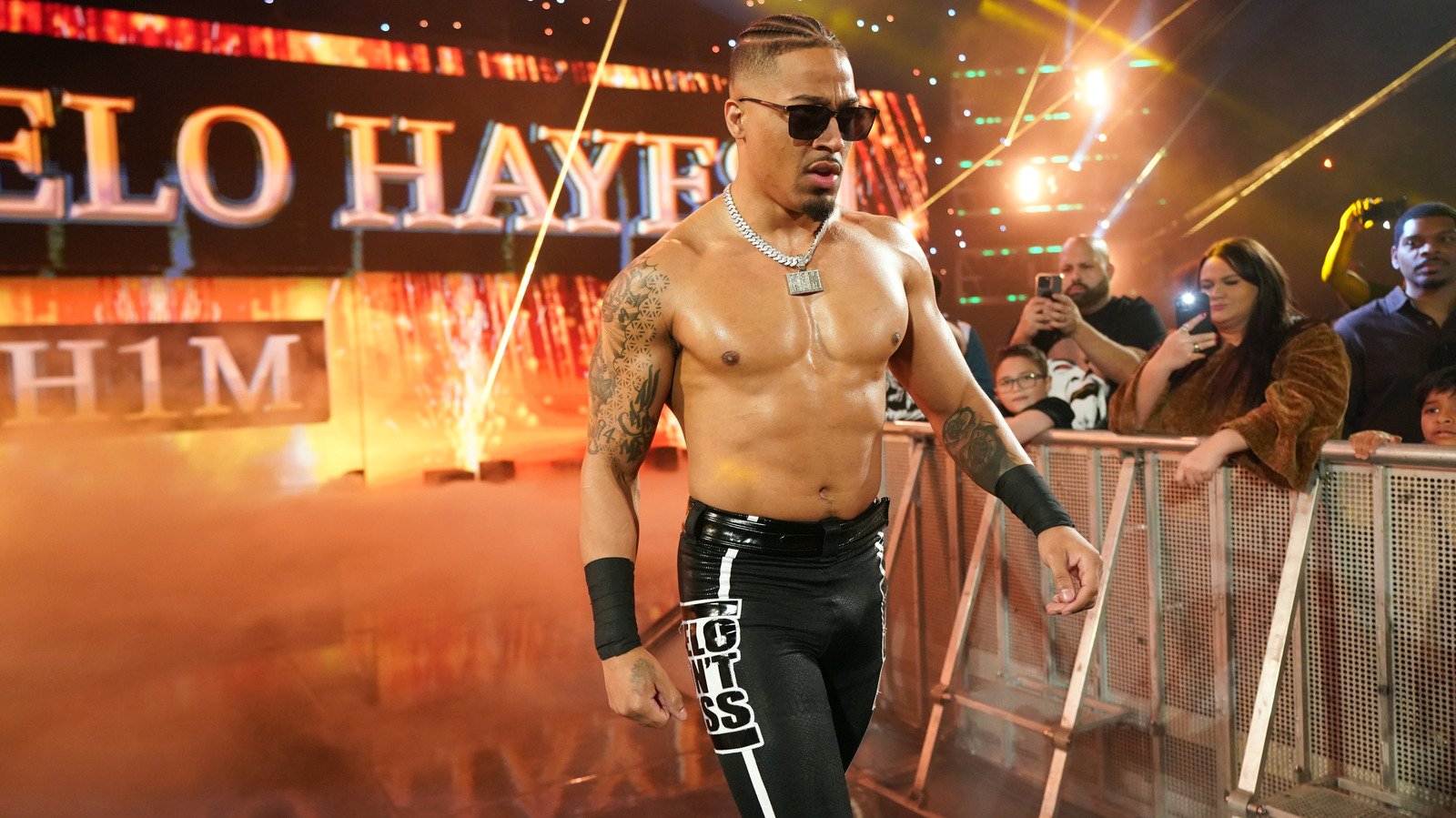Professional wrestler Carmelo Hayes walks down a ramp in an arena with fire effects behind him, wearing black tights and sunglasses, while fans in the background take photos.