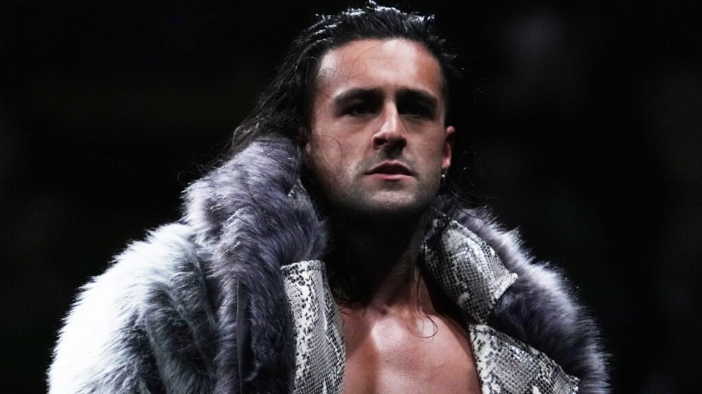 NJPW wrestler Drilla Moloney wearing a grey fur-lined snakeskin jacket and looking intensely toward the camera.