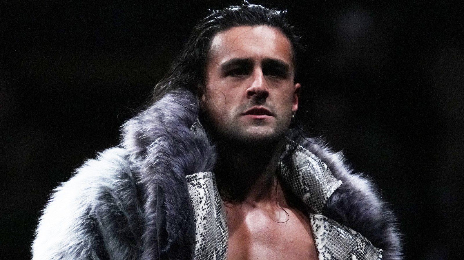 NJPW wrestler Drilla Moloney wearing a grey fur-lined snakeskin jacket and looking intensely toward the camera.
