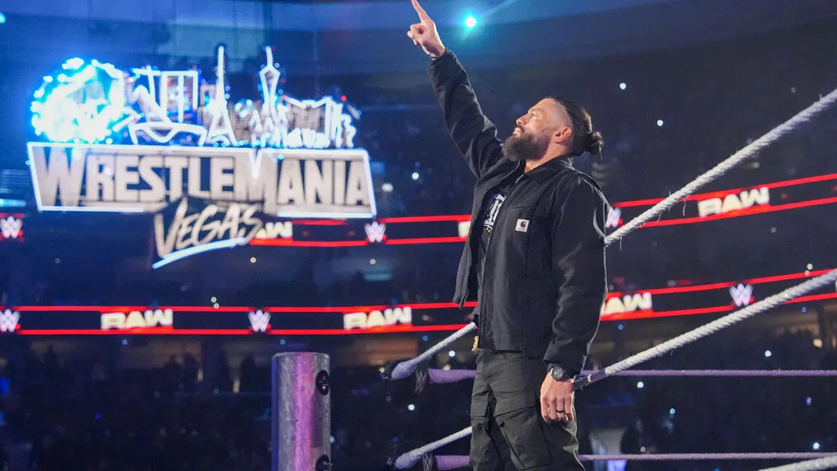 Professional wrestler Roman Reigns stands in a WWE ring, pointing a single finger upward toward a large "WrestleMania Vegas" sign displayed on the arena's screen.