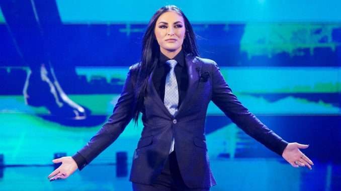 Professional wrestler Sonya Deville, wearing a black suit and a light grey tie, poses with her arms outstretched in a blue-lit wrestling ring.