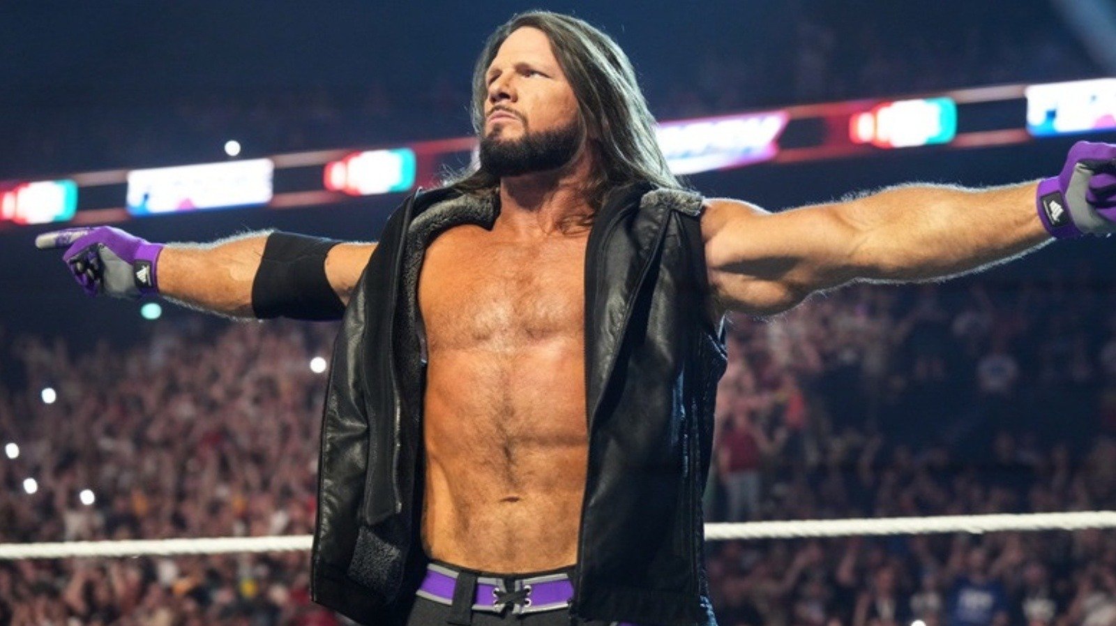 Professional wrestler AJ Styles, a man with long brown hair and a goatee, smiles while spreading his arms wide in a wrestling ring.