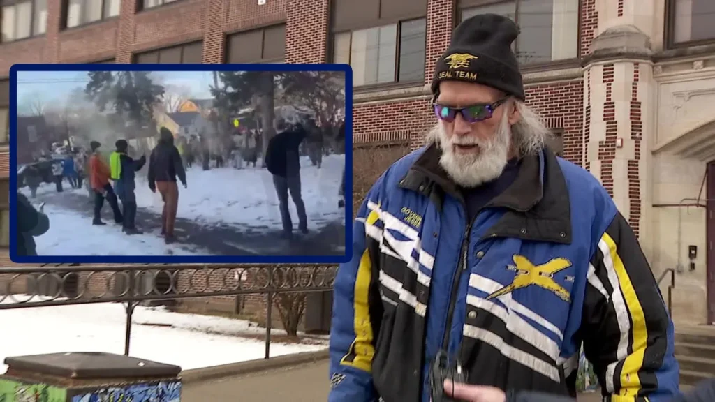 Jesse Ventura, a man with a gray beard and "SEAL Team" beanie, speaks outdoors at a high school, while an inset box shows a snowy scene of people gathered.