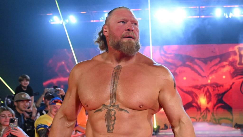 The beast incarnate returns: brock lesnar declares for the 2026 royal rumble - tjr wrestling A close-up, high-intensity photograph of a shirtless, heavily muscled brock lesnar standing on a wwe entrance ramp. He has a serious expression, looking off to the side with blonde hair tied back in a small ponytail and a light beard. A large, prominent tattoo of a vertical sword with a brass-knuckle hilt is visible in the center of his chest. Bright yellow laser lights and stadium spotlights illuminate him from behind against a backdrop of a fiery, skull-like graphic on an led screen and a cheering crowd.