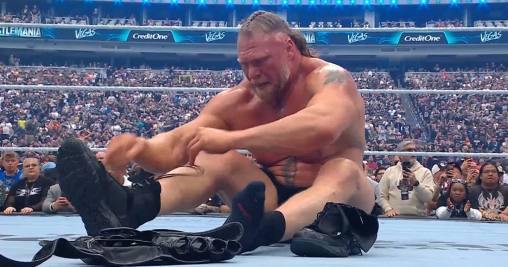 A wide shot of a wrestling ring at allegiant stadium during wrestlemania 42. Brock lesnar, a massive, muscular man with a blonde braided mohawk and goatee, is sitting on the blue ring canvas. He is shirtless and wearing black wrestling trunks. He is in the process of unlacing and removing his black wrestling boots, having already placed his signature mma-style gloves on the mat in front of him. In the blurred background, thousands of fans in the stadium stands are watching the moment, with "vegas" and "wrestlemania" signage visible on the arena tiers.