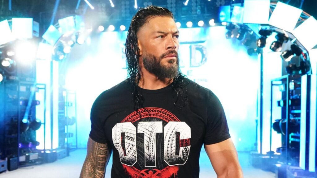 Wwe superstar roman reigns, with long wet-look dark hair and a full beard, stands in a wrestling arena under bright blue and white stage lights. He is wearing a black t-shirt with the bold white letters "otc" across the chest. He has a serious expression, looking off to the side as he stands in the center of the entrance ramp. - tjr wrestling