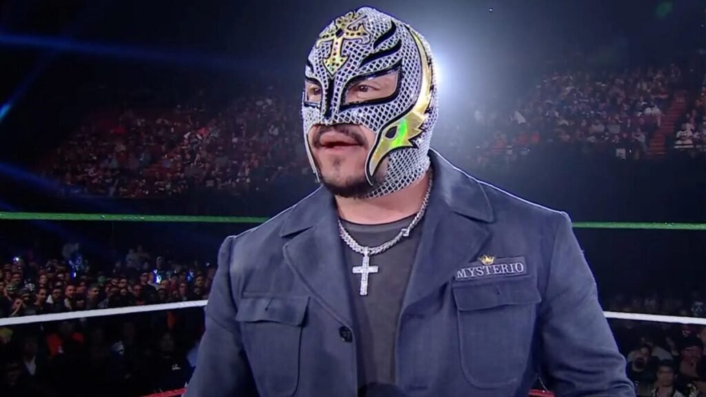 A medium shot of wwe hall of famer rey mysterio standing in a wrestling ring. He is wearing a silver and gold lucha libre mask with a cross on the forehead and a greyish-blue button-up "lwo" jacket that has "mysterio" and a gold crown embroidered on the left chest. He is looking off-camera with a neutral expression. The background shows a large, blurred crowd in a dark arena with red seating and green ring ropes. - tjr wrestling