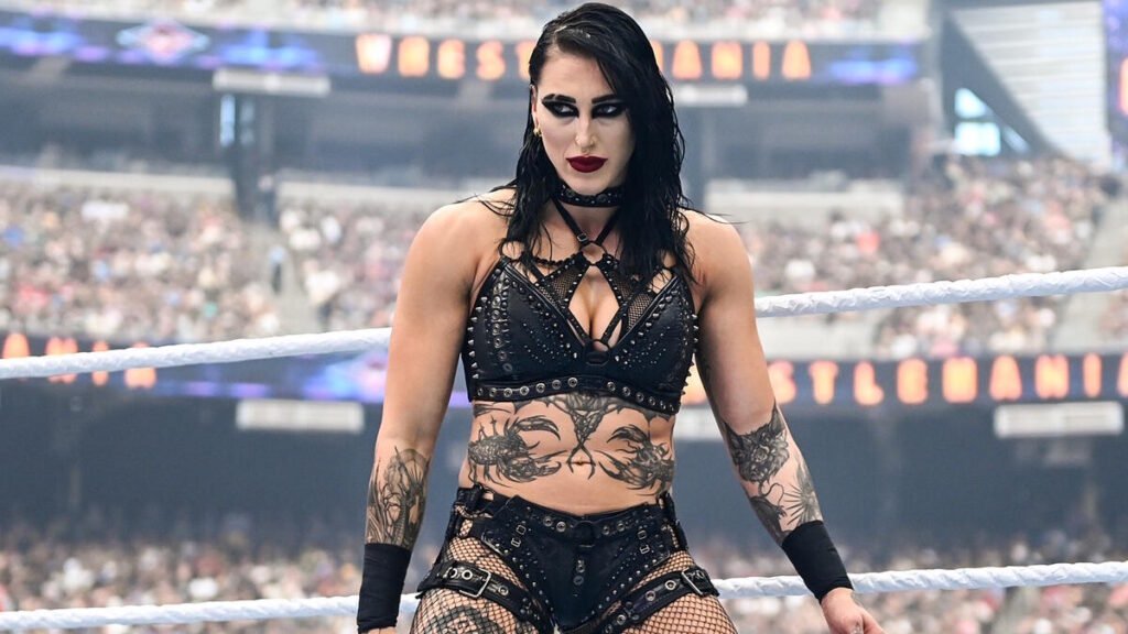 Rhea ripley stands inside a wwe ring with an intense expression, wearing black ring gear and visible tattoos, as a packed stadium crowd fills the background.