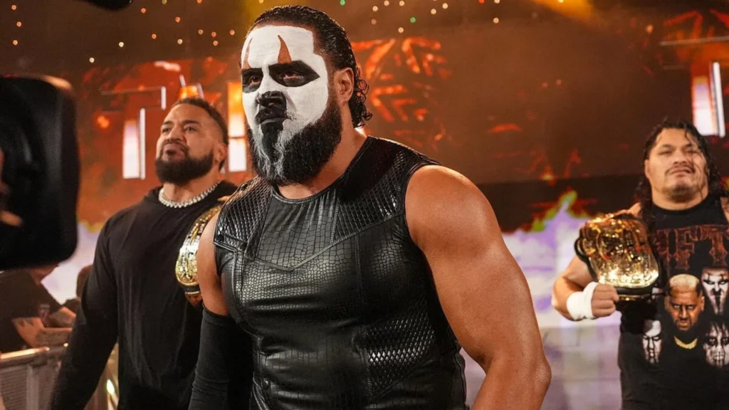 Professional wrestlers jacob fatu, tama tonga, and tanga loa stand together in a dimly lit wwe arena. Fatu, in the center, wears striking white and black face paint and a textured black sleeveless vest, looking forward with a menacing expression. To his left, tama tonga looks off-camera with a serious gaze. On the right, tanga loa holds a gold championship belt over his shoulder. The background features warm, orange-toned stage lighting and subtle pyrotechnic sparks.