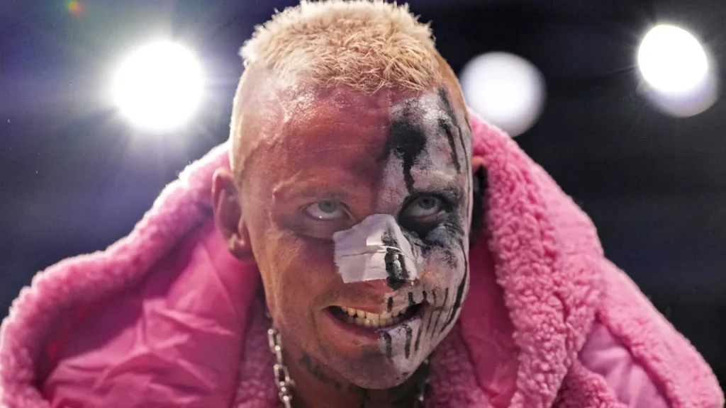 Darby allin stares intensely at the camera with black and white face paint, a taped nose, blonde hair, and a pink fluffy jacket during an aew event.