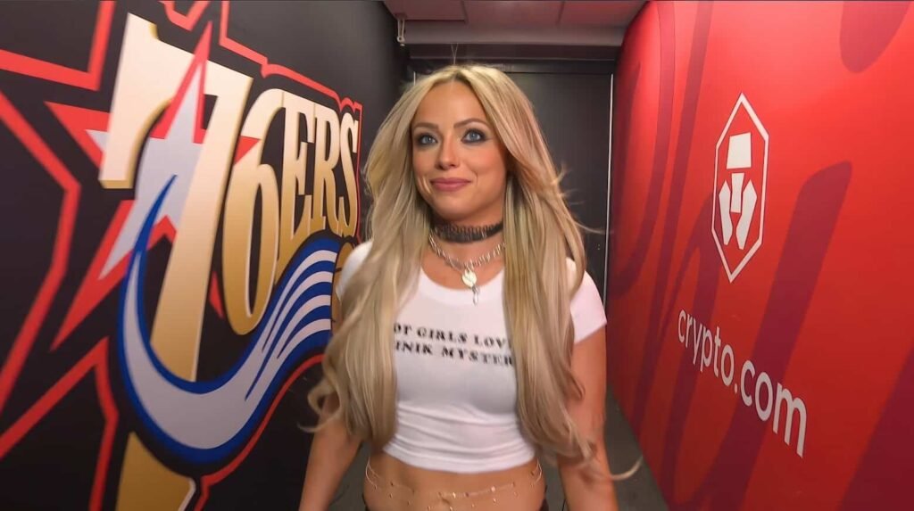 Liv morgan backstage appearance on wwe show - tjr wrestling Liv morgan standing backstage in a wwe arena hallway, smiling while wearing a white top and choker, with branding visible on the walls.