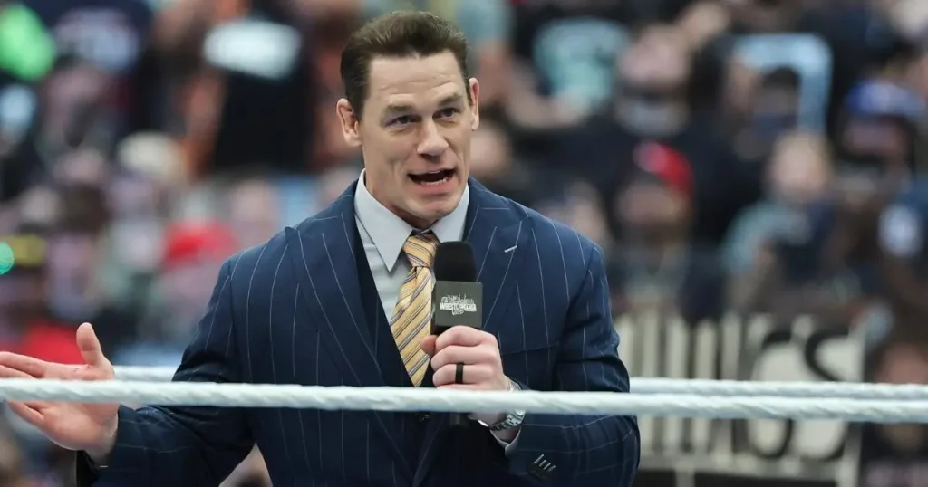 A medium shot of wwe superstar john cena standing in a wrestling ring. He is wearing a sophisticated navy blue pinstripe suit with a matching vest, a white dress shirt, and a gold-and-navy diagonal striped tie. He holds a black wwe-branded microphone in his right hand, gesturing with his left as he speaks with an intense, passionate expression. The background is a large, blurred arena crowd, and a white ring rope is visible in the foreground across his chest. - tjr wrestling