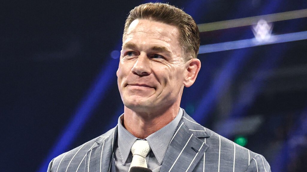 A close-up shot of professional wrestler and actor john cena. He is looking off-camera with a warm, slightly emotional smile. He is wearing a blue and white pinstriped suit with a crisp white tie. His hair is neatly styled in a short, modern fade. The background is a soft-focus arena with scattered blue and green lights, suggesting a large-scale event. - tjr wrestling
