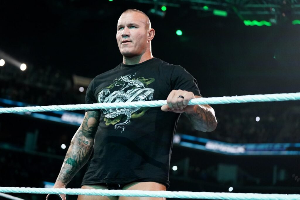 Randy orton stands inside the wwe ring, gripping the ropes with one hand while wearing a black graphic t-shirt, looking intensely toward the crowd.