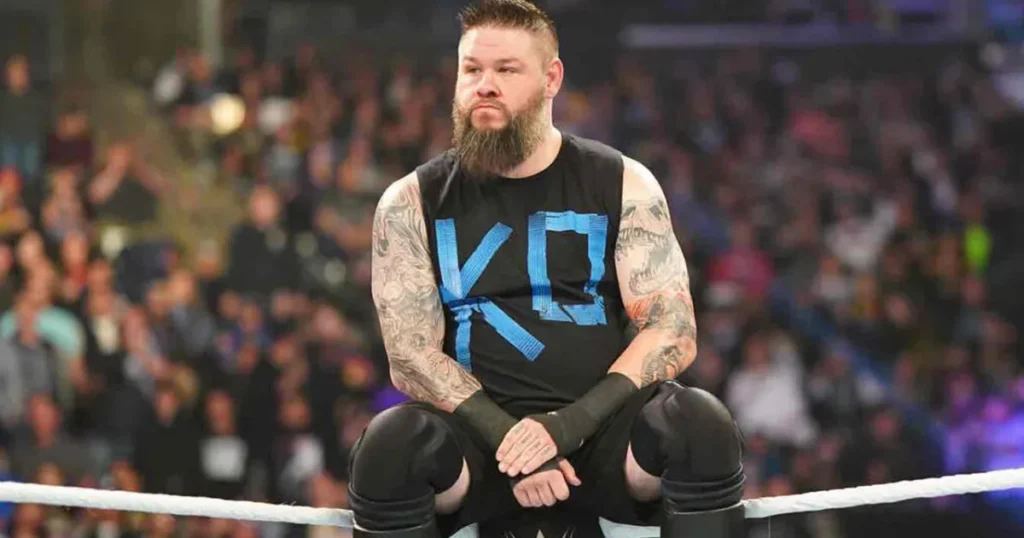 Kevin owens sits on the ring apron during a wwe event, wearing a black ko shirt and looking focused toward the crowd.