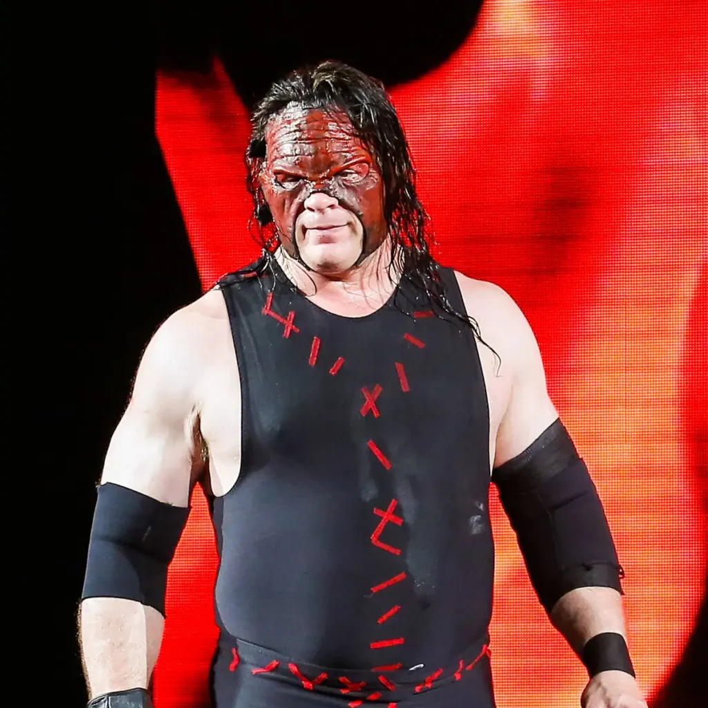 A large, muscular wwe wrestler wearing a black mask, black singlet with red slash markings, and black gloves stands inside a wrestling ring with arena crowd and lights visible in the background. - tjr wrestling