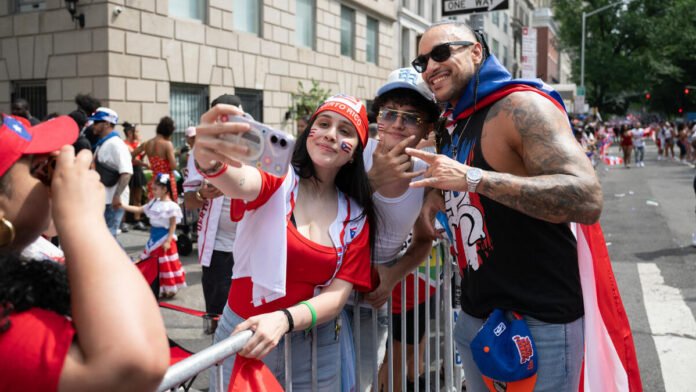 A young woman with a Puerto Rican flag painted on her cheek takes a selfie with a group of friends at a street parade, including a heavily tattooed man wearing a Puerto Rican cape and sunglasses, with crowds of parade-goers dressed in red, white, and blue lining the barricaded street behind them.
