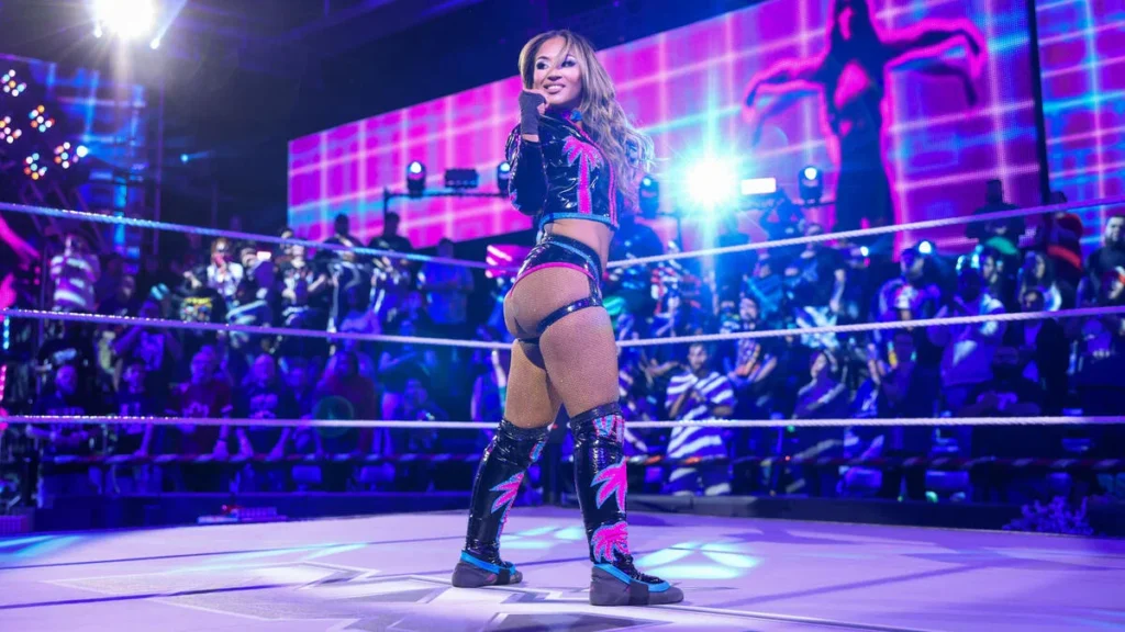Lola vice poses in the ring during wwe nxt appearance - tjr wrestling Lola vice standing in a wwe ring, glancing back over her shoulder with a smile, wearing black and pink ring gear including a crop jacket, shorts, fishnet stockings, and tall black boots with pink palm tree designs, in front of a vibrant pink and purple led arena backdrop and a live crowd.