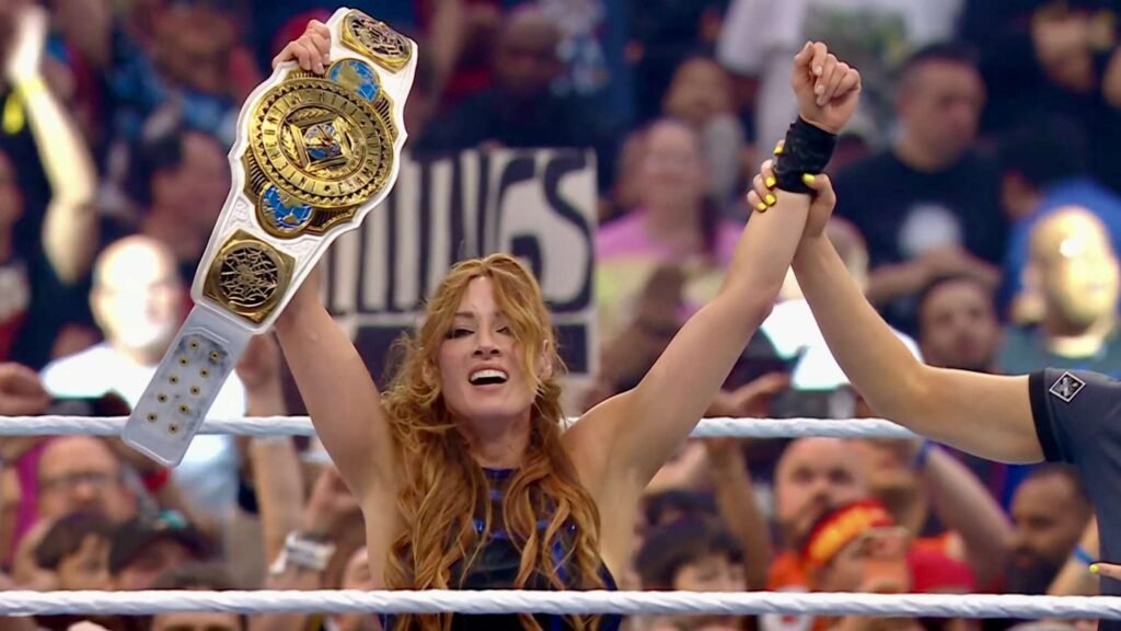 A female professional wrestler raises both arms in victory inside a wrestling ring, holding a white and gold championship belt aloft in her left hand while a referee raises her right hand. She has long, wavy auburn hair, wears a blue and black ring gear, and has yellow-painted nails. A large crowd fills the arena in the background. - tjr wrestling