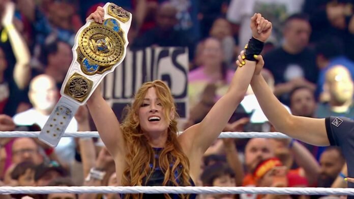 A female professional wrestler raises both arms in victory inside a wrestling ring, holding a white and gold championship belt aloft in her left hand while a referee raises her right hand. She has long, wavy auburn hair, wears a blue and black ring gear, and has yellow-painted nails. A large crowd fills the arena in the background.
