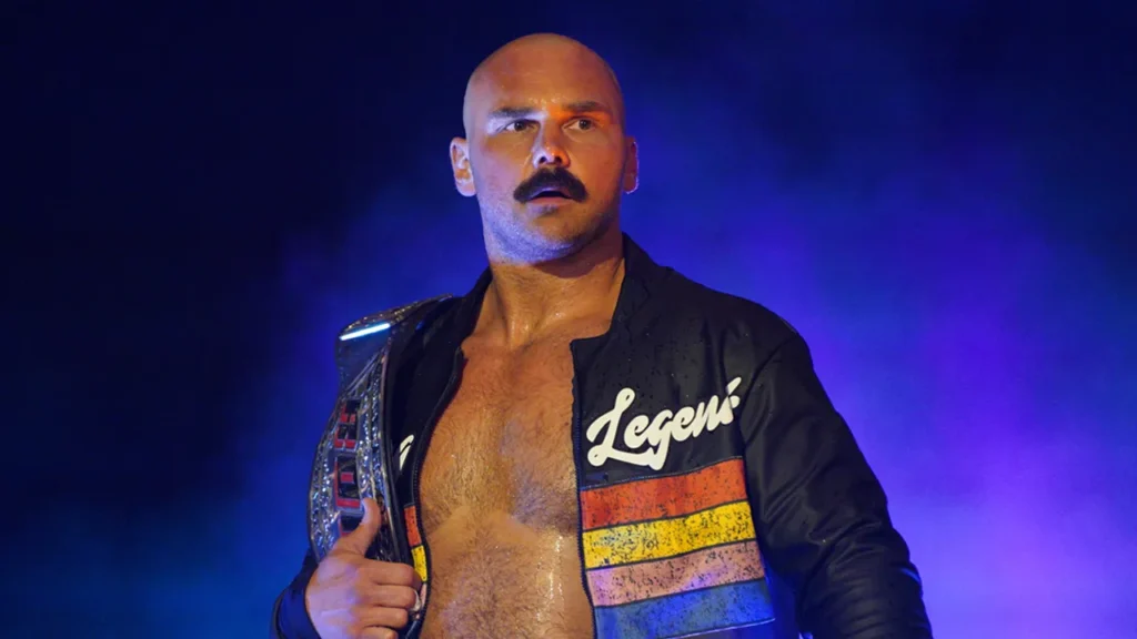 Dax harwood, a bald professional wrestler with a thick mustache, standing against a blue and purple smoky backdrop while holding a championship belt over his shoulder, wearing an open black leather jacket with rainbow stripes and "legends" written in script across the chest.