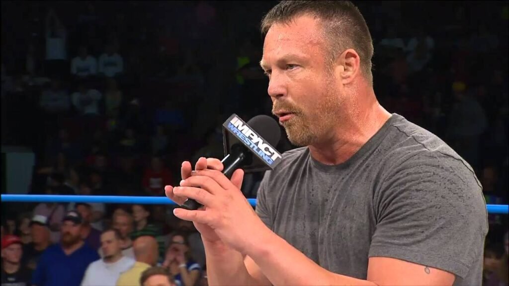 A muscular man in a grey t-shirt speaking into an impact wrestling branded microphone inside a wrestling ring, with a packed arena crowd visible in the blurred background. - tjr wrestling