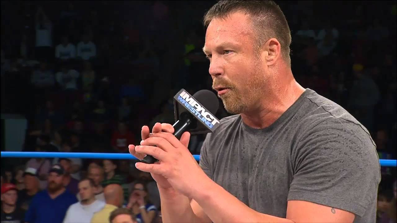 A muscular man in a grey t-shirt speaking into an impact wrestling branded microphone inside a wrestling ring, with a packed arena crowd visible in the blurred background. - tjr wrestling