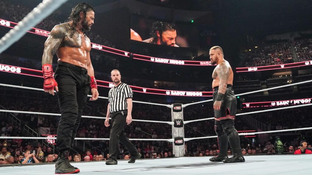 Roman reigns, heavily tattooed and shirtless in black pants and red fingerless gloves, stares down a blonde-haired opponent in black and red ring gear inside a wwe ring, with a referee in a black and white striped shirt standing between them, a packed arena crowd and large video board visible in the background during a wwe raw event.