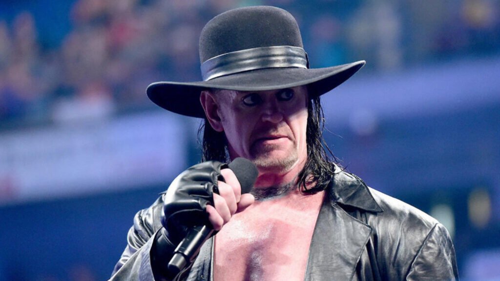 The undertaker addresses the wwe universe with chilling in-ring promo - tjr wrestling A wwe wrestler dressed as the undertaker character stands in the ring holding a microphone close to his mouth with a black leather gloved hand. He wears a wide-brimmed black hat, has long dark hair, a goatee, and is dressed in an open black leather coat with a bare chest. His expression is intense and menacing. A blurred arena crowd is visible in the blue-lit background.