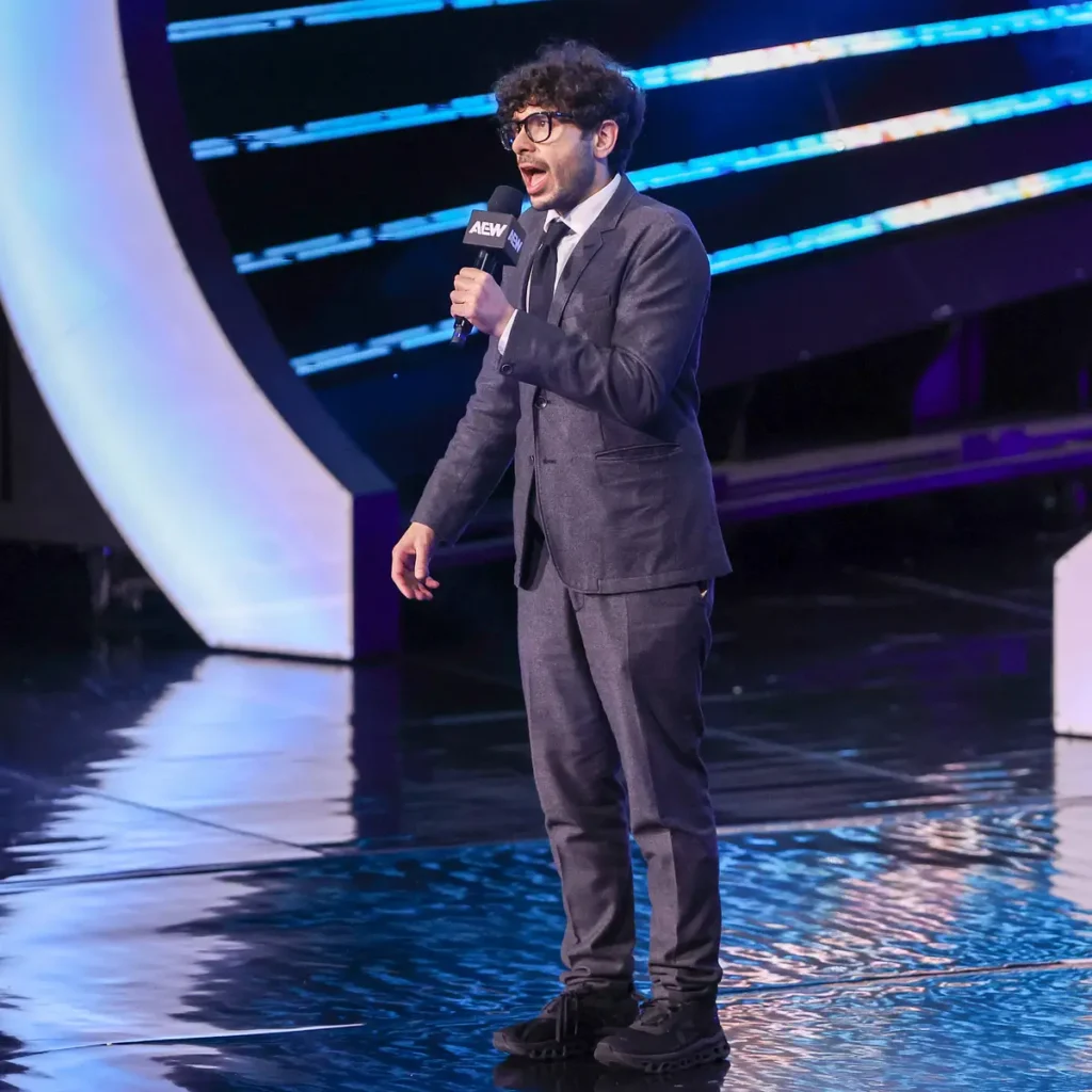 Aew president tony khan addressing the crowd - tjr wrestling Aew president tony khan wearing a dark suit, tie, and glasses while speaking into an aew-branded microphone on stage during a live show.