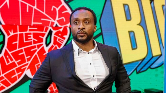 WWE Superstar Big E stands in front of a colorful LED screen wearing a formal dark suit jacket and white shirt.