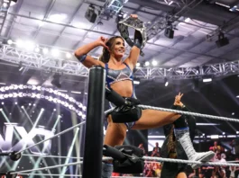 WWE NXT superstar Kendal Grey poses atop the ring ropes in blue and silver ring gear, raising the NXT Women's Championship belt in celebration.