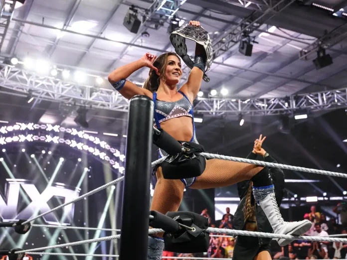 WWE NXT superstar Kendal Grey poses atop the ring ropes in blue and silver ring gear, raising the NXT Women's Championship belt in celebration.